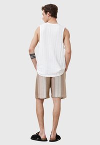 Weißes ärmelloses Shirt aus strukturiertem Stoff, kombiniert mit braunen gestreiften Shorts. Schwarze Sandalen runden das Outfit ab. Ansicht von hinten.