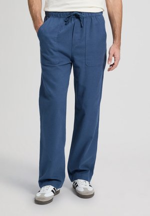 Mann trägt blaue Freizeithose mit Kordelzug und weiße Sneaker mit schwarzen Streifen, steht mit entspannten Händen, eine Hand in der Tasche.
