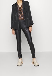 Blazer noir à double boutonnage sur un top blouson fleuri, associé à des leggings noirs brillants et des chaussures à plateformes beiges.