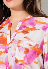 Blusa de botones en blanco con un estampado de acuarela que presenta tonos de rosa, naranja y marrón; incluye un pequeño bolsillo en el pecho y un cuello.