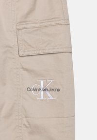 Pantalons cargo en coton beige avec une poche latérale. Caractéristiques d'un logo brodé blanc "Calvin Klein Jeans" et "CK." Détails de couture subtils.