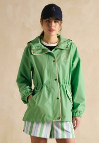 Zaļa, ūdensnecaurlaidīga jaka ar auklu jostas daļai, zelta pogām un baltu apdari. Apvienota ar svītrainām šortiem, ar kapuci un kabatām.