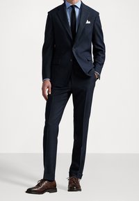 Mies, joka on pukeutunut tummansiniseen pukuun, jossa on lahkeet taitettuna, vaaleansiniseen kauluspaitaan, tummaan solmioon, valkoiseen taskuliinaan, ruskeisiin nahkakenkiin ja rannekelloon.