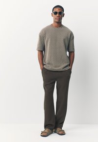 Tan textured short-sleeve shirt with diamond pattern, paired with brown trousers and green sandals. Accessories include dark sunglasses.