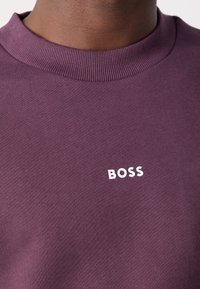 Gros plan sur un cou à la peau foncée portant un t-shirt violet à col rond avec le texte blanc « BOSS » sur la poitrine.