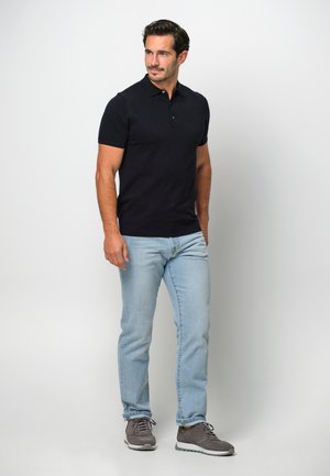 Marineblaues Poloshirt mit kurzen Ärmeln und einer Knopfleiste mit drei Knöpfen, kombiniert mit hellblauen Jeans und grauen Sneakers, stehend vor einem weißen Hintergrund.