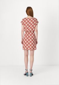 Robe rouge à motifs floraux, avec des manches courtes et une taille nouée. Associée à des sandales à talons blocs bleu. Fond blanc uni.