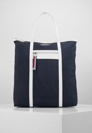 Navy blue tote bag with white straps, front zipper pocket, Tommy logo near top, and striped zipper pull detail, standing on a white surface.
