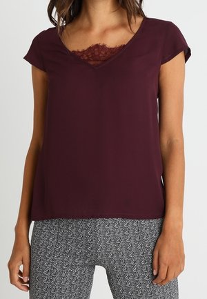 Femme portant un haut bordeaux foncé à manches courtes avec une bordure en dentelle au niveau du col en V, associé à un pantalon à motif chevrons noir et blanc.