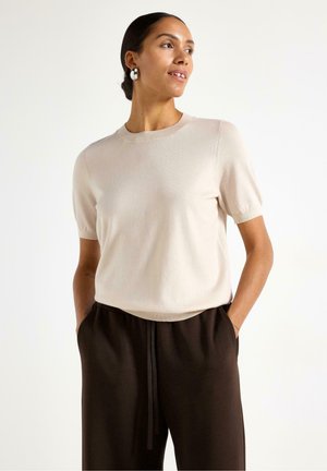 Beige strikket kortærmet top med rund halsudskæring. Fremstillet af glat stof med en diskret tekstur, kombineret med løstsiddende mørkebrune bukser.