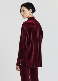 Camicetta in velluto bordeaux con maniche lunghe, una piega sul retro e polsini decorati. Il tessuto ha una consistenza morbida e una finitura lucida.