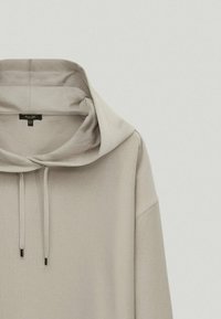 Light beige hoodie with a large, draped hood, drawstrings, and relaxed fit. Soft fabric, minimal seams, and a tag visible near the neckline.