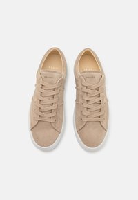 Sneakers in suede beige con suole in gomma bianca, dotate di lacci piatti e logo dorato sulla linguetta. Design pulito e minimalista.
