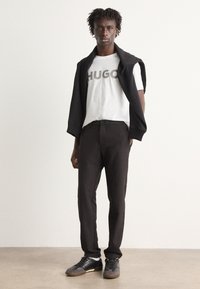 Black sweater draped over shoulders, white T-shirt with "HUGO" text, black trousers, and black sneakers. Smooth fabric, slim fit design.