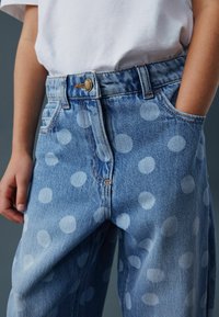 Blugi din denim albastru deschis cu buline mari albe, având închidere cu nasturi și buzunare frontale. Materialul are o textură ușor decolorată.