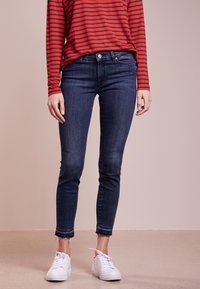 Mörkblå skinny jeans med fransade fållar, i kombination med en långärmad topp i röd och svart randig design samt vita sneakers. Golvet är ljust.