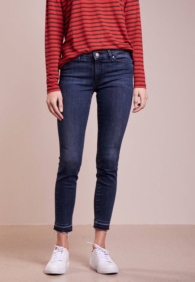 Mörkblå skinny jeans med fransade fållar, i kombination med en långärmad topp i röd och svart randig design samt vita sneakers. Golvet är ljust.