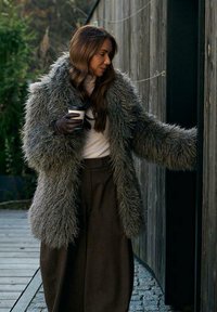 Kobieta ubrana w szary płaszcz z sztucznego futra i brązową spódnicę, trzymająca kubek kawy, sięgająca, by otworzyć drewniane drzwi na zewnątrz.