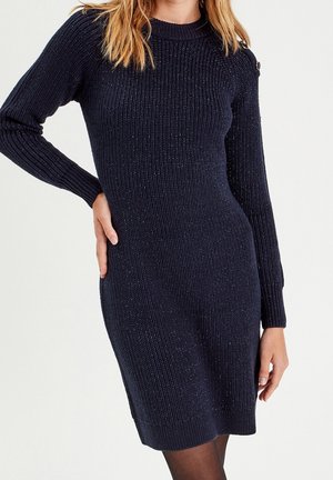 Femme portant une robe-pull à manches longues en maille côtelée bleu marine foncé avec des boutons sur les épaules, posant avec une main sur la taille.
