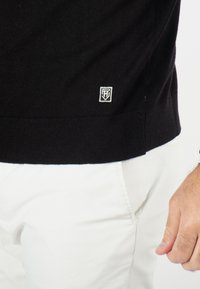 Pull en tricot noir avec un ourlet côtelé, arborant un petit logo blanc sur le bord inférieur. Porté avec un pantalon blanc, montrant une texture lisse.