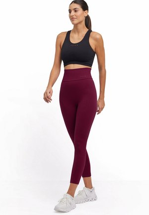 Vrouw loopt, gekleed in een zwarte sportbeha, high-waisted getextureerde bordeauxrode legging en witte sportschoenen tegen een witte achtergrond.