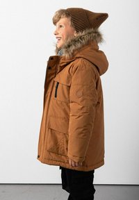 Brązowa kurtka puchowa z kapturem wykończonym futrem, zamek błyskawiczny i zapięcia na zatrzaski, liczne kieszenie oraz brązowa czapka typu beanie o ściśniętym wzorze na uśmiechniętym dziecku na jednolitym tle.