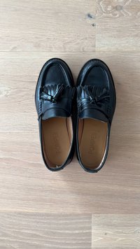 Melna ādas loaferu pāris ar pušķiem un franjām, ar uzrakstu "POMPEII," novietoti blakus uz gaišas koka grīdas.