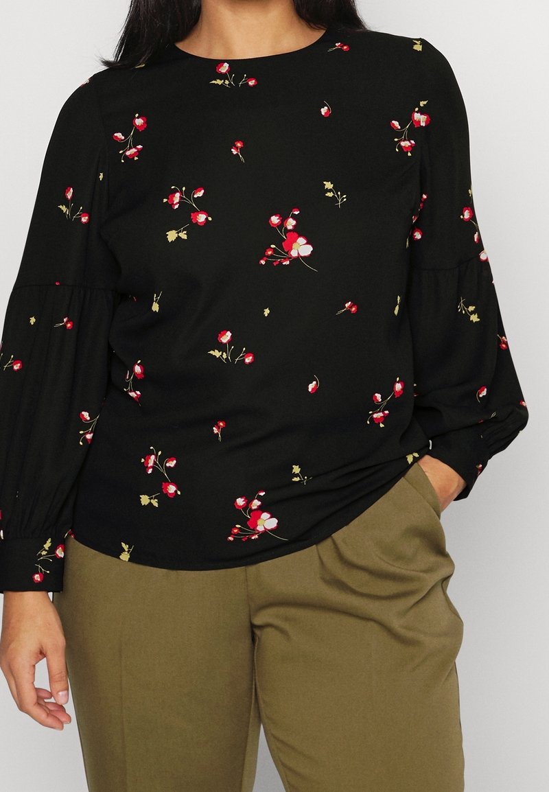 Kvinde i sort langærmet bluse med små røde og hvide blomsterprint, parret med olivengrønne bukser, den ene hånd i lommen.