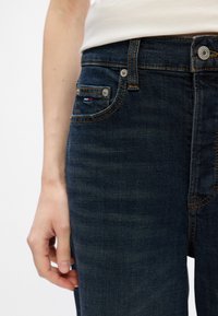 Sötétkék farmernadrág, slim fit szabással, sárga cérnával varrva, elülső zsebével, amely piros és fehér varrással készült, és fémből készült gombbal a deréknál.
