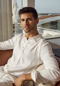 Camicia bianca in lino con una texture sottile, dotata di colletto e maniche arrotolate, indossata da un uomo seduto su un divano all'interno.