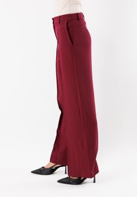 Pantaloni larghi bordeaux realizzati in tessuto morbido; presentano pieghe, tasche laterali e una vita sagomata. Abbinati a tacchi a spillo neri.
