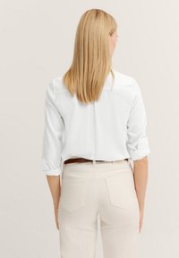Weißes, langärmliges Hemd mit hochgekrempelten Ärmeln und Rückenfalten, kombiniert mit hellen Hosen, die ein sauberes, einfaches Design zeigen.