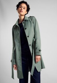 Ljusgrön trenchcoat med en slät textur, dubbelknäppt design, svarta knappar och åtsittande midja, i kombination med mörka jeans.