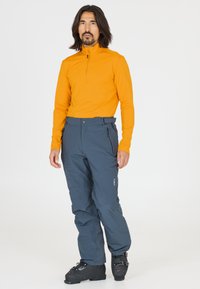 Orange langærmet bluse med lynlås ved halsen; mørkegrå, løstsiddende skibukser med sidelommer; sorte skistøvler med spænder og tekstureret overflade.