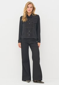 Juoda mygtukais užsegama marškinėliai su krūtinės kišenėmis, kontrastine siūle ir ilgomis rankovėmis, derinami su juodomis plačiomis kelnėmis ir smeigtukais puoštomis basutėmis.