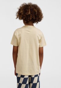 Beige bomulds t-shirt med korte ærmer og rund halsudskæring, vist fra bagsiden. Kombineret med marineblå og cremefarvet ternede bukser.