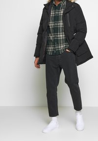 Zwarte pufferjack met hood van nepbont, geruite groene overhemden, donkergrijze jeans en witte sneakers. Geweven stof en casual pasvorm.