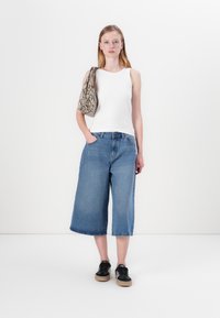 Hvit ermeløs topp, vide denim culotter i lyseblå, svarte sneakers og en teksturert beige veske med slangeskinnmønster.