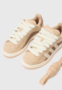 Beige suede sneakers with cream thick laces, featuring light brown accents and three stripes on the side. Includes an extra rolled shoelace.