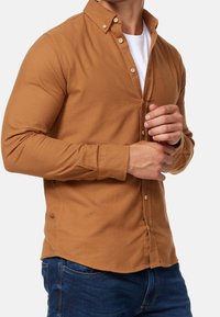 Braune Hemdbluse aus weichem Stoff, mit klassischem Kragen, langen Ärmeln und geradem Saum, getragen über einem weißen T-Shirt.