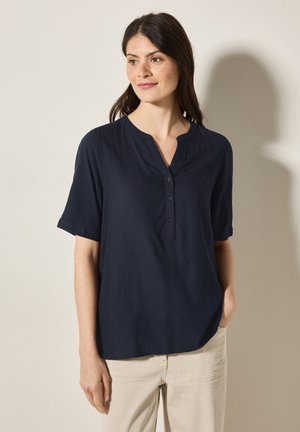 Blouse bleu marine à manches courtes avec un col mandarin, fermeture à boutons et coupe décontractée. Tissu lisse avec un subtil éclat, associée à un pantalon clair.