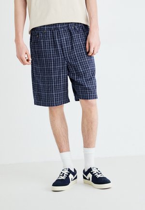 Mand iført marineblåternede shorts, hvide sokker og marineblå sneakers står mod en simpel hvid baggrund.