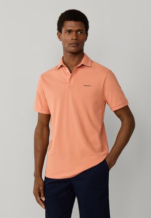 Hombre con camisa tipo polo color salmón con el logo "Hackett" y pantalones azul marino, de pie con una mano en el bolsillo contra un fondo liso.