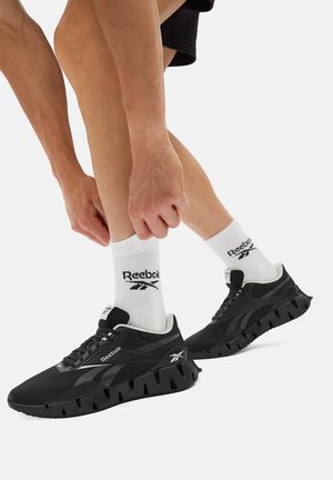 Tenisi negri Reebok au un superior texturat, o talpă groasă cu caneluri și detalii subtile cu logo. Purtați cu șosete albe care afișează logo-ul.
