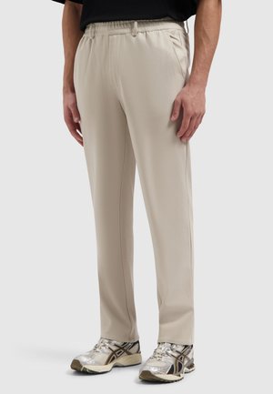 Man draagt beige rechte broekspijpen en zilver-zwarte hardloopschoenen, staat met ontspannen handen langs zijn zij tegen een lichte achtergrond.