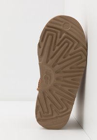 Semelle d’une botte UGG marron avec un motif de bande de roulement en zigzag, positionnée debout contre un fond blanc d’angle.
