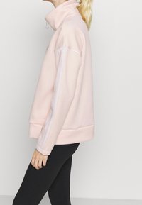 Sweat-shirt rose à col montant avec poignets côtelés, doté de bandes blanches le long des manches et d'un tissu texturé. Associé à des leggings noirs.