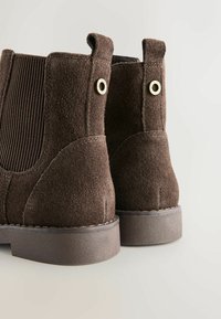 Botas de tobillo de ante marrón con lados de tejido acanalado, costuras en contraste y acentos de hardware dorado en la parte trasera. Suelas de goma bajas.
