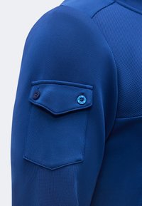 Gros plan d'une manche de chemise bleue à manches longues avec une poche à rabat boutonnée comportant un bouton bleu étiqueté "SEVE."