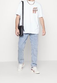 Vit grafisk t-shirt med ett färgglatt tryck, ljusblå denimjeans och vita sneakers med röda accenter. Svart crossbody-väska.
